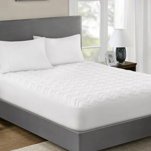 A bed with white fitted sheet, two pillows, and a quilted mattress pad, beside a nightstand with a lamp.
