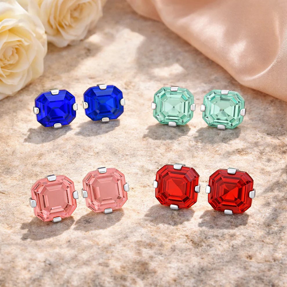Four pairs of square-cut gemstone stud earrings in blue, green, pink, and red, set in metal clasps, displayed on a stone surface with roses and fabric in the background.