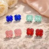 Four pairs of square-cut gemstone stud earrings in blue, green, pink, and red, set in metal clasps, displayed on a stone surface with roses and fabric in the background.