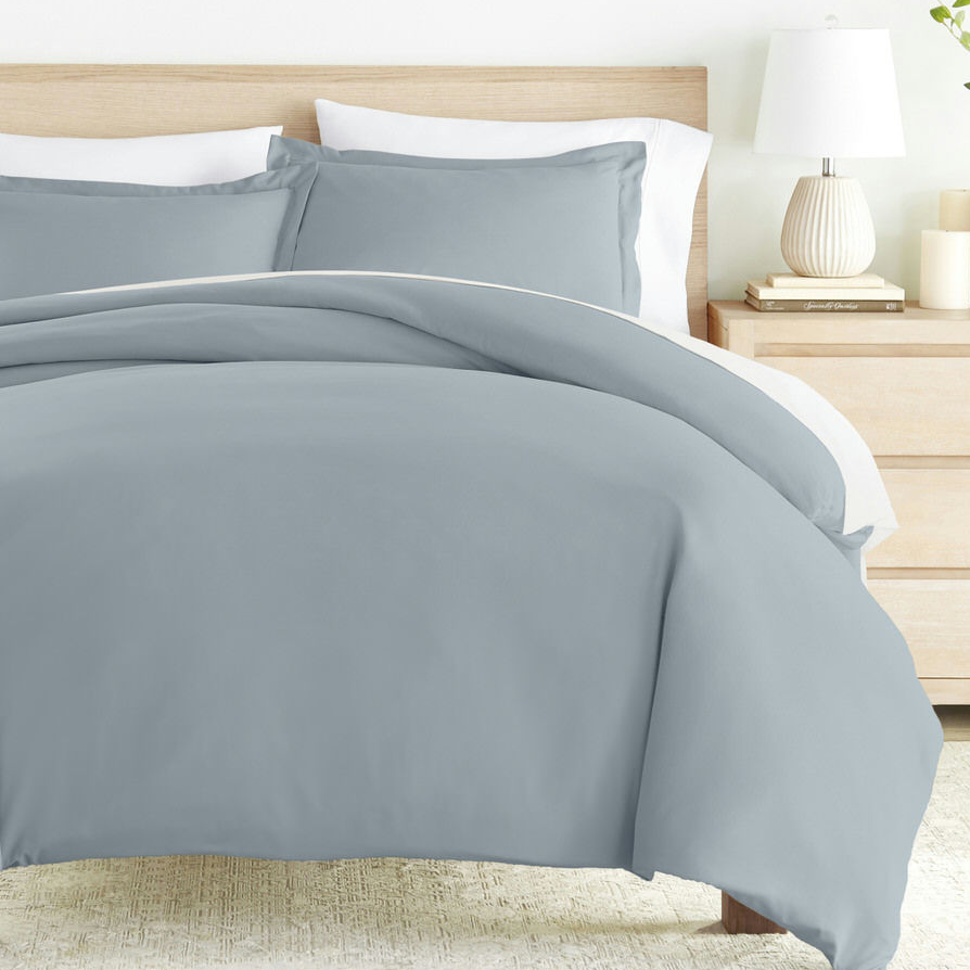 A neatly arranged bed with light gray bedding, including a duvet cover and pillows, set against a wooden headboard. A bedside table with a lamp and a stack of books is visible.