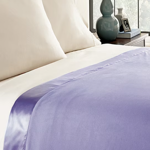 Lightweight Queen Flannel Blanket in lavender with a smooth satin edge, draped over a bed with white pillows. The scene includes a bedside table with a decorative vase, photo frame, and other small items near a window.