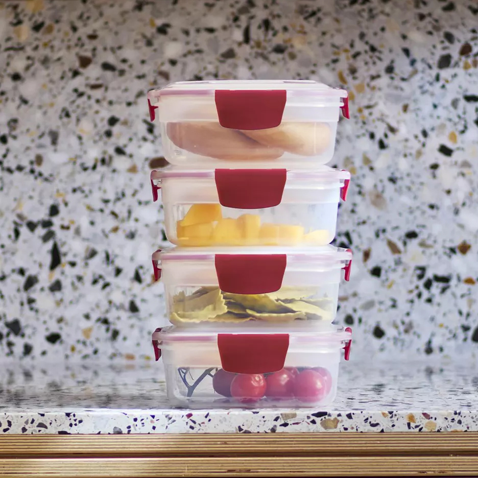 Stack of four food storage containers with red clips, containing various foods: meat, sliced mango, pasta, and tomatoes, against a terrazzo countertop background.