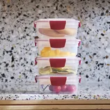 Stack of four food storage containers with red clips, containing various foods: meat, sliced mango, pasta, and tomatoes, against a terrazzo countertop background.