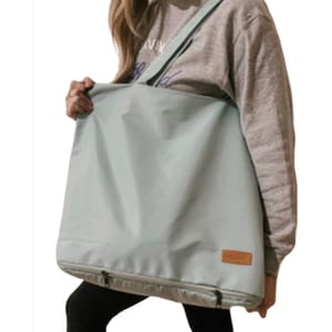 A person carrying a large, light blue tote bag with a shoulder strap and a small leather patch. The bag has a zipper at the bottom.