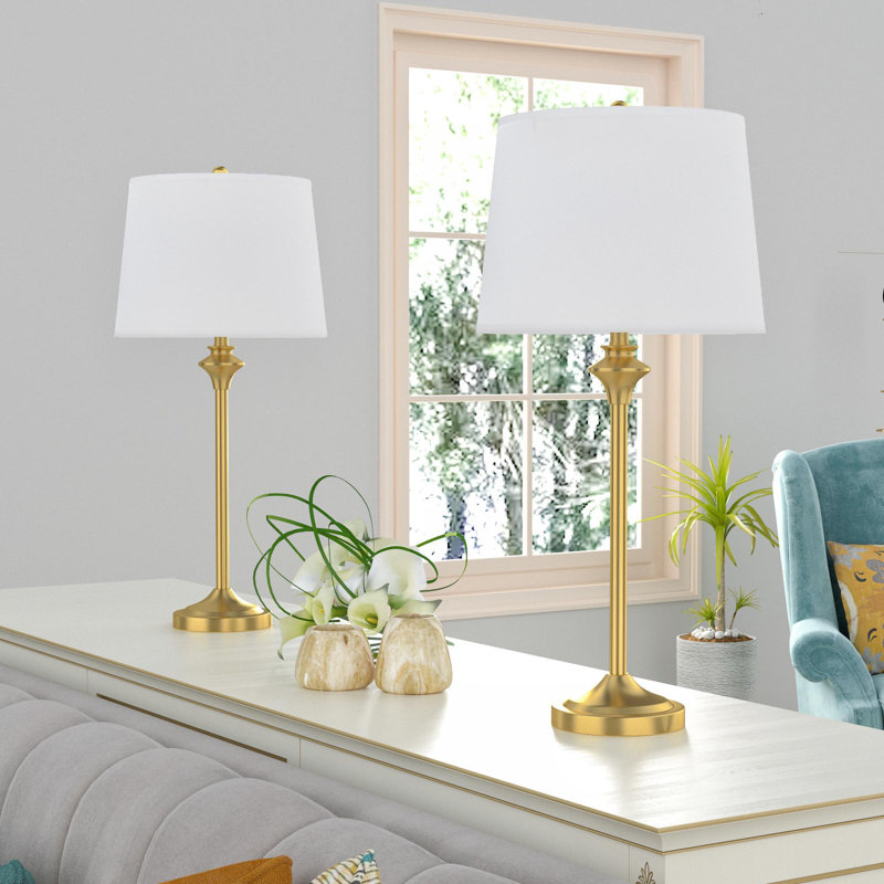 Two matching gold table lamps with white shades on a white sideboard, accompanied by decorative flower vases and potted plants.