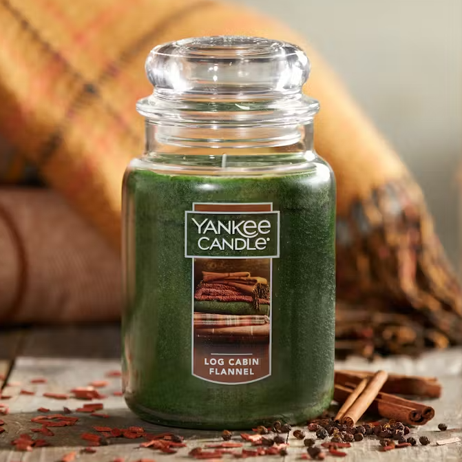 A green Yankee Candle with a \“Log Cabin Flannel\“ label, accompanied by a picture of stacked flannel blankets, stands next to cinnamon sticks and cloves.