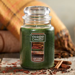 A green Yankee Candle with a \“Log Cabin Flannel\“ label, accompanied by a picture of stacked flannel blankets, stands next to cinnamon sticks and cloves.