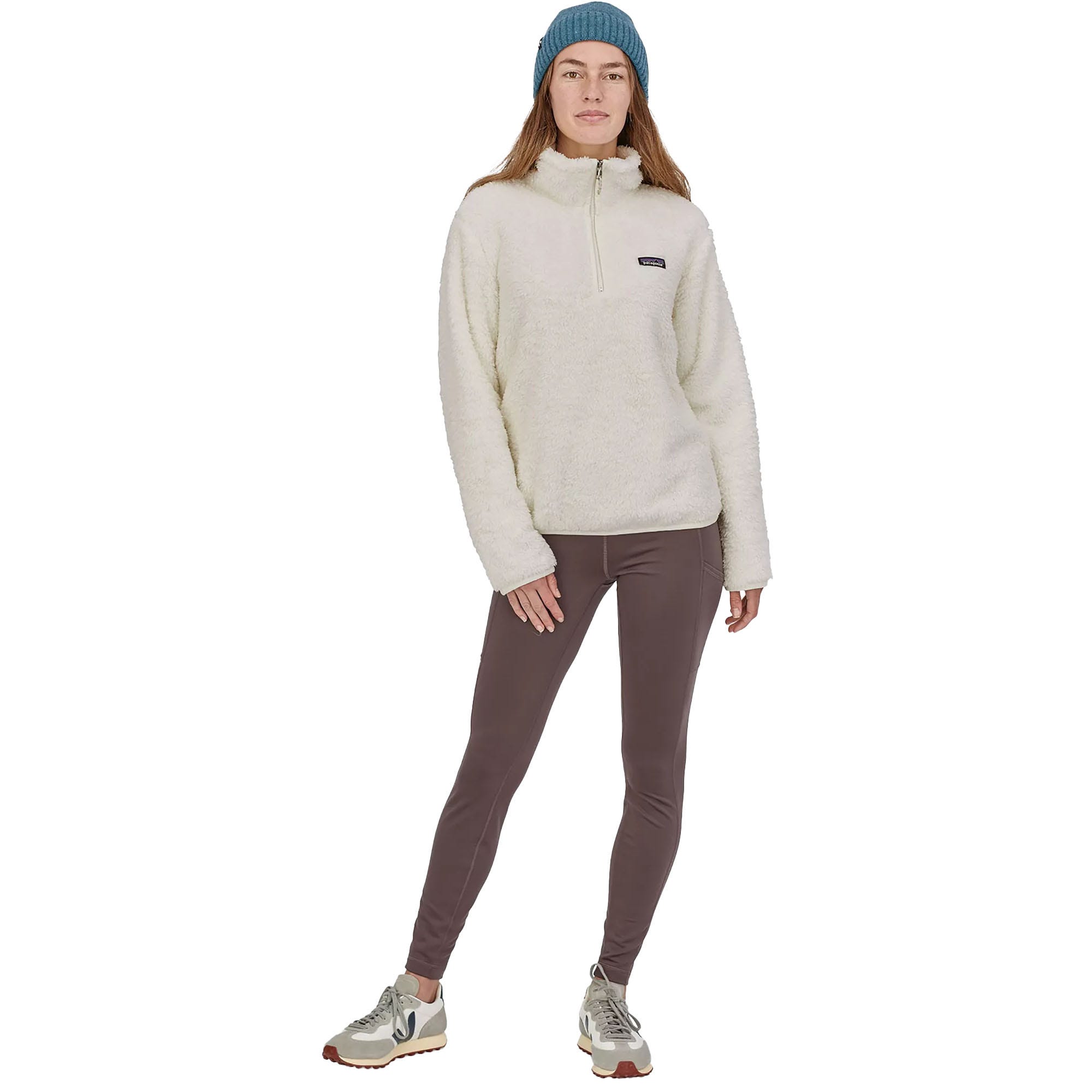 A woman is wearing a white fleece pullover, brown leggings, beige sneakers, and a blue beanie.