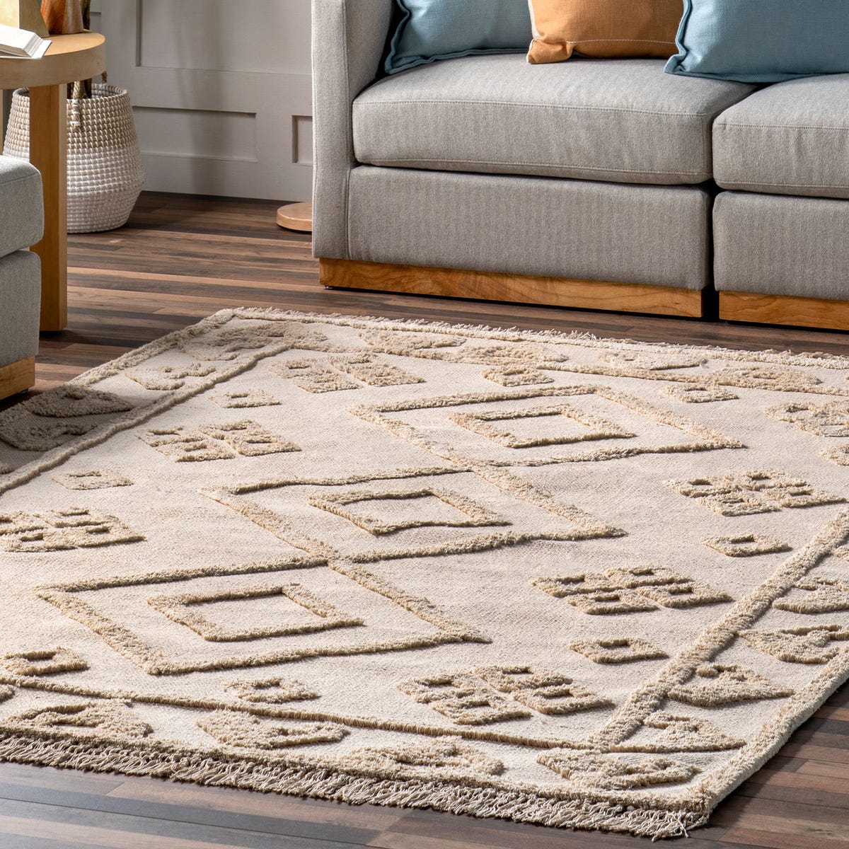 A beige area rug with a geometric pattern is placed on a wooden floor, next to a gray sofa with colorful cushions.