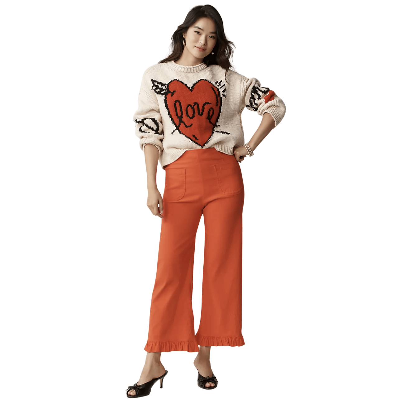 A woman is wearing a cream sweater with a red heart and \“love\“ design paired with high-waisted orange pants featuring a ruffled hem, and black heeled sandals.