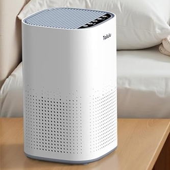 A white air purifier with a perforated design on the lower half and control buttons on the top, placed on a wooden surface with a bed in the background.