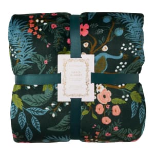 A folded fleece blanket features a dark background with a colorful floral and botanical pattern, including various flowers and leaves, held together by a dark green ribbon.