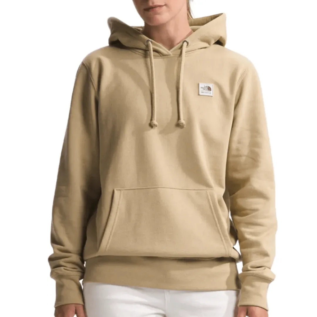 The North Face Heritage Hoodie is a beige pullover with a kangaroo pocket, a drawstring hood, and a small logo patch on the chest. It offers a casual and comfortable fit.