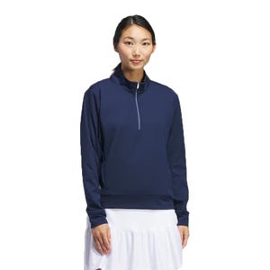 Adidas Ultimate Layering Pullover features a navy-colored, long-sleeved design with a quarter-zip collar. It has a relaxed fit suitable for layering, and is paired with a white skirt in the image.
