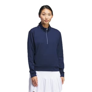Adidas Ultimate Layering Pullover features a navy-colored, long-sleeved design with a quarter-zip collar. It has a relaxed fit suitable for layering, and is paired with a white skirt in the image.