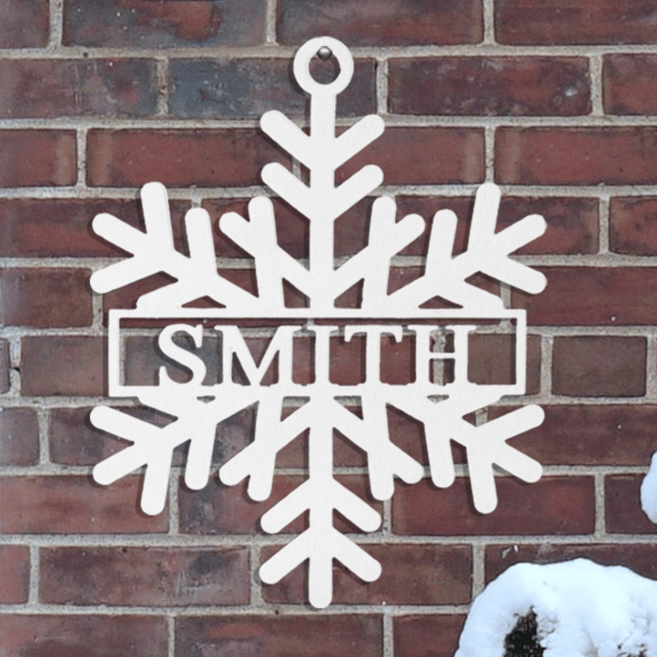 A snowflake-shaped wall hanging features the word \“SMITH\“ in the center, mounted on a brick wall beside a snow-covered bush.