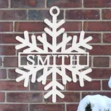 A snowflake-shaped wall hanging features the word \“SMITH\“ in the center, mounted on a brick wall beside a snow-covered bush.