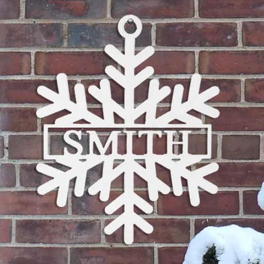 A snowflake-shaped wall hanging features the word \“SMITH\“ in the center, mounted on a brick wall beside a snow-covered bush.
