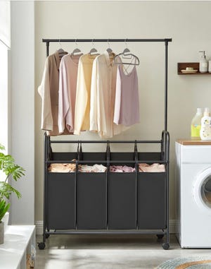 A laundry cart with four black fabric bins and a hanging rod above, set on a metal frame with wheels, is shown in a room with a washing machine and home decor.