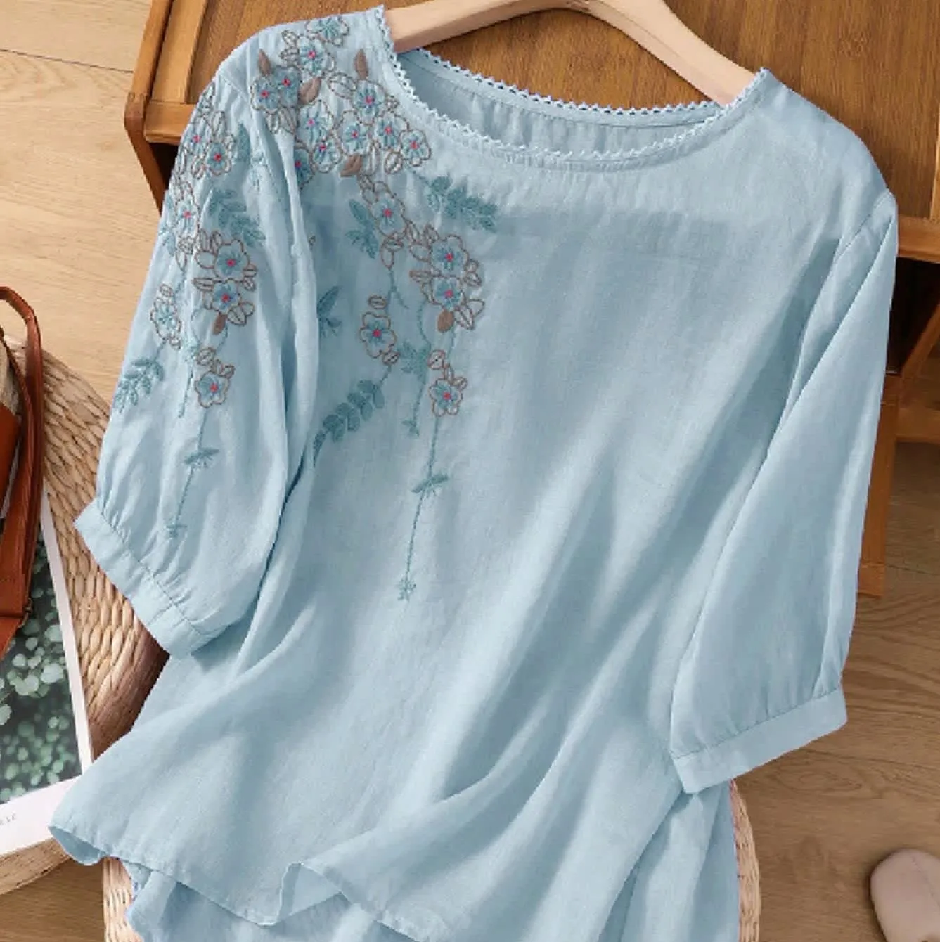 Light blue blouse with floral embroidery on the shoulder and sleeves, featuring a round neck and three-quarter length sleeves.