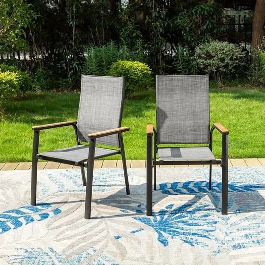 Two modern outdoor chairs with gray mesh fabric and wooden armrests are placed on a patterned rug, set against a green lawn and lush garden background.