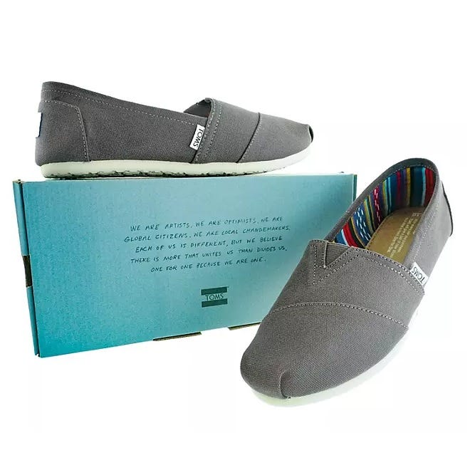 A pair of gray TOMS slip-on shoes with a colorful striped lining is displayed with a branded shoebox featuring a message about global citizenship and unity.