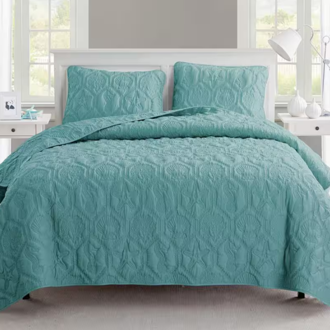 A teal quilt with a textured, seashell and starfish pattern is displayed on a bed with matching pillow shams. The setting includes light-colored bedside tables and decor, creating a serene bedroom environment.