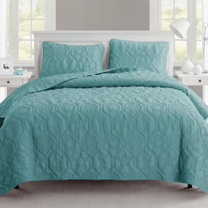 A teal quilt with a textured, seashell and starfish pattern is displayed on a bed with matching pillow shams. The setting includes light-colored bedside tables and decor, creating a serene bedroom environment.