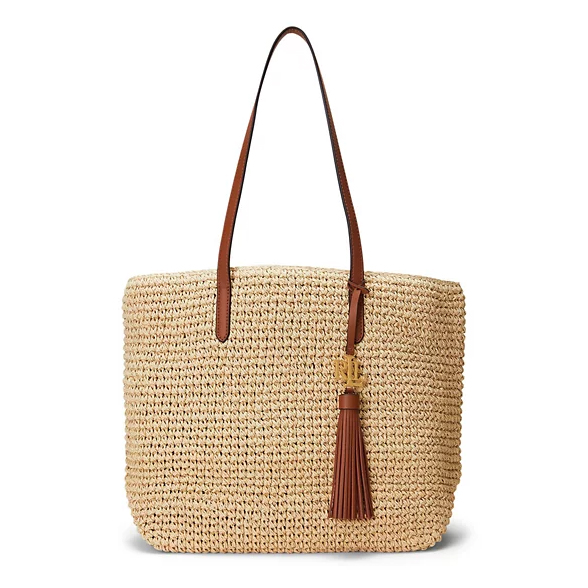 A straw tote bag with brown leather handles and a decorative tassel, featuring a gold monogram charm.
