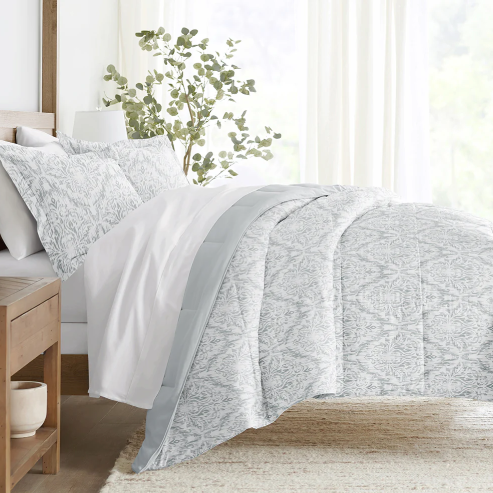 A neatly made bed features a light gray patterned comforter and pillow shams, with a plain white sheet. A wooden nightstand and a plant in the background complete the serene bedroom setting.