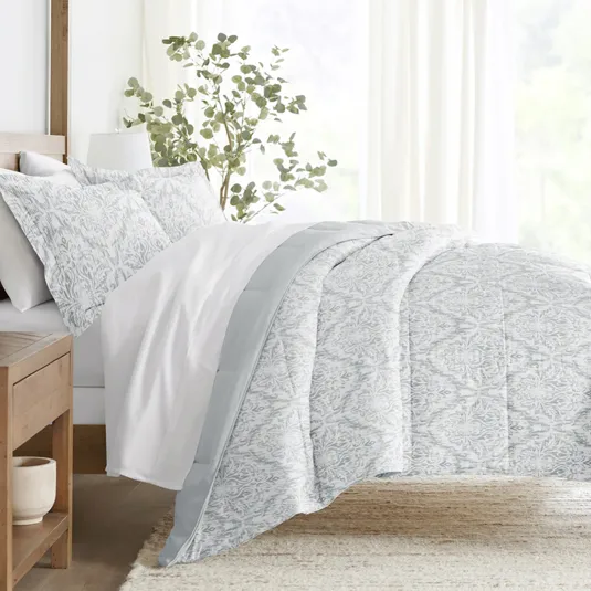 A neatly made bed features a light gray patterned comforter and pillow shams, with a plain white sheet. A wooden nightstand and a plant in the background complete the serene bedroom setting.