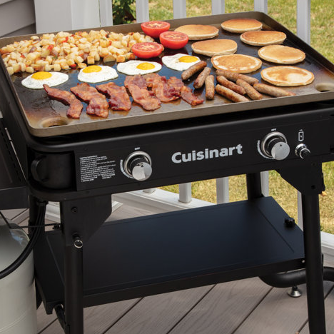 A Cuisinart griddle cooking a variety of breakfast items, including eggs, bacon, pancakes, sausages, tomatoes, and potatoes, on a deck.