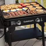 A Cuisinart griddle cooking a variety of breakfast items, including eggs, bacon, pancakes, sausages, tomatoes, and potatoes, on a deck.