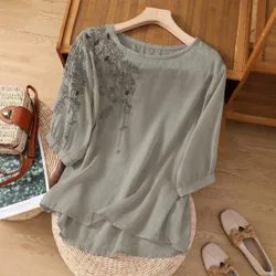 A light grey blouse with floral embroidery on the shoulders and sleeves is displayed on a hanger. Nearby are a woven handbag with a brown strap and a pair of beige loafers with bows.