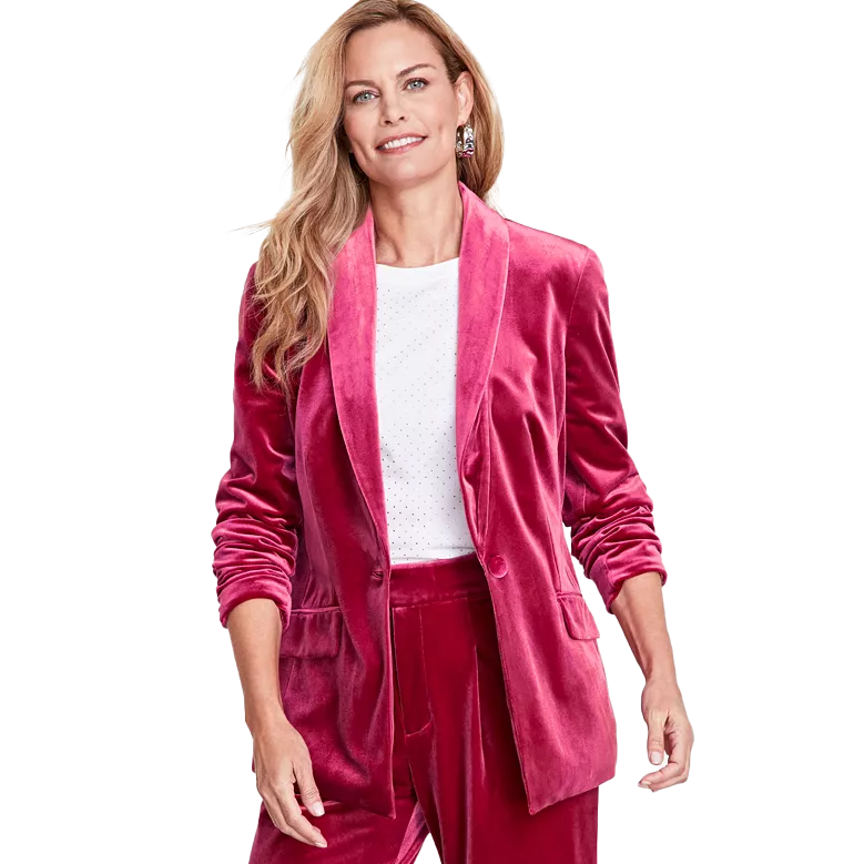 A woman models a vibrant pink velvet blazer with a shawl collar and long sleeves over a dotted white shirt. The outfit is completed with matching pants, giving a festive or holiday appearance.