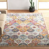 A brightly colored area rug with intricate geometric patterns is placed on a wooden floor in a living room. A beige sofa with a striped pillow is on the left, a wicker basket with magazines is in the corner, and a cactus plant is next to a wall with a cow portrait.