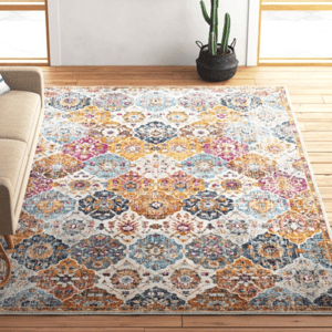 A brightly colored area rug with intricate geometric patterns is placed on a wooden floor in a living room. A beige sofa with a striped pillow is on the left, a wicker basket with magazines is in the corner, and a cactus plant is next to a wall with a cow portrait.