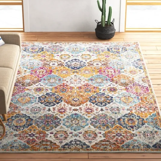 A brightly colored area rug with intricate geometric patterns is placed on a wooden floor in a living room. A beige sofa with a striped pillow is on the left, a wicker basket with magazines is in the corner, and a cactus plant is next to a wall with a cow portrait.