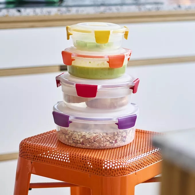 Stack of round, clear plastic food containers with colored locking lids, placed on an orange stool.