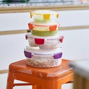 Stack of round, clear plastic food containers with colored locking lids, placed on an orange stool.