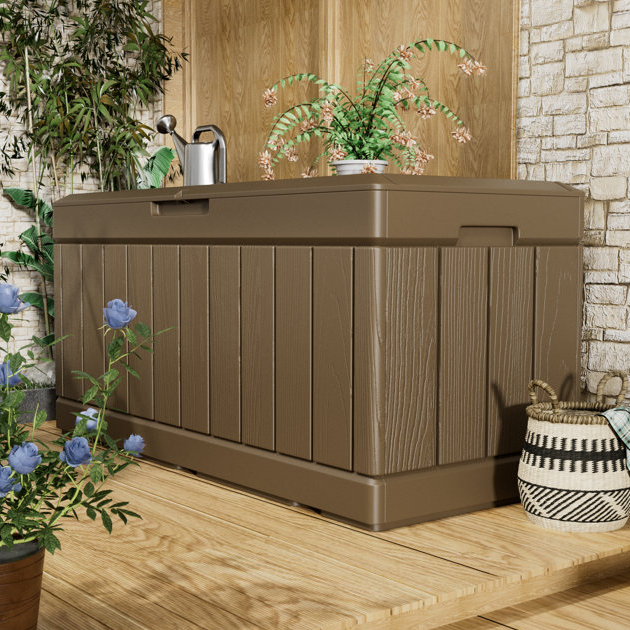 A large brown outdoor storage box with a wood-like texture is placed on a wooden deck, surrounded by potted plants and a wicker basket.