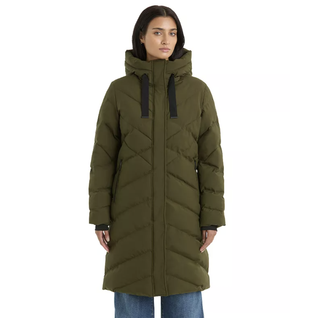 A woman is wearing an olive green, long puffer coat with a hood and black drawstrings, paired with blue jeans.