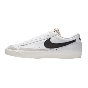White low-top Nike Blazer Vintage shoes feature a black swoosh, beige suede accents on the toe, and a white rubber sole. The heel displays \“NIKE\“ in black text.