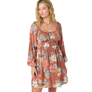 A woman is wearing a floral-patterned, long-sleeved dress with a square neckline in warm, earthy tones.