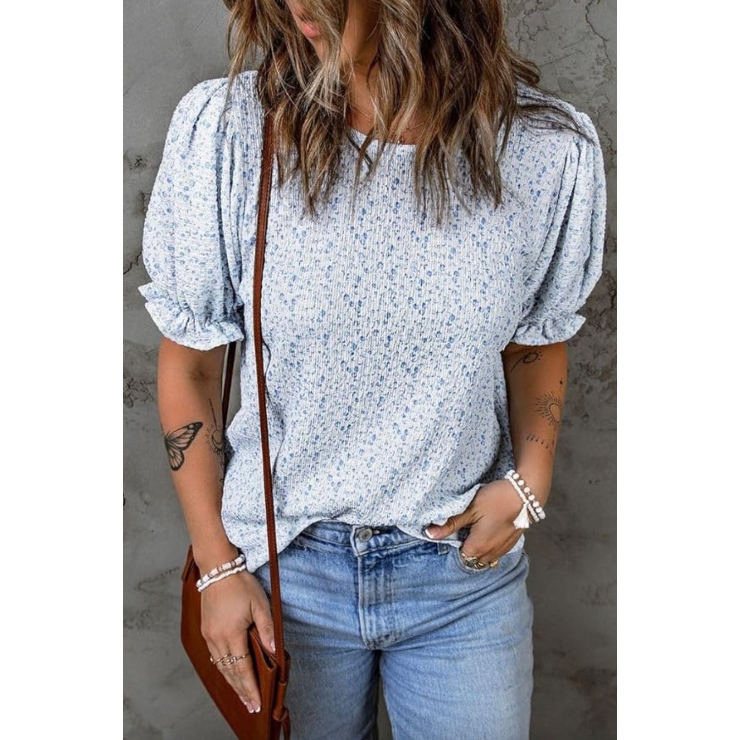 A woman is wearing a floral print blouse with puff sleeves and blue denim jeans, accessorized with bracelets and a crossbody bag.