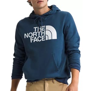 The North Face Half Dome Hoodie is a blue pullover with a drawstring hood, featuring the brand's logo prominently in white on the chest and a front kangaroo pocket. It is paired with beige pants.
