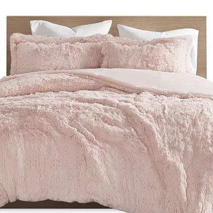 A bed with a light pink, fluffy comforter set, including matching pillow shams and a natural wood headboard.