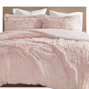A bed with a light pink, fluffy comforter set, including matching pillow shams and a natural wood headboard.