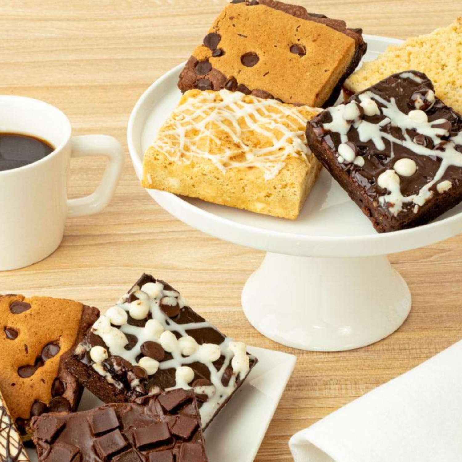 A variety of brownies and blondies with different toppings, such as chocolate chips and drizzled icing, are displayed on a cake stand and plate alongside a cup of black coffee on a wooden surface.