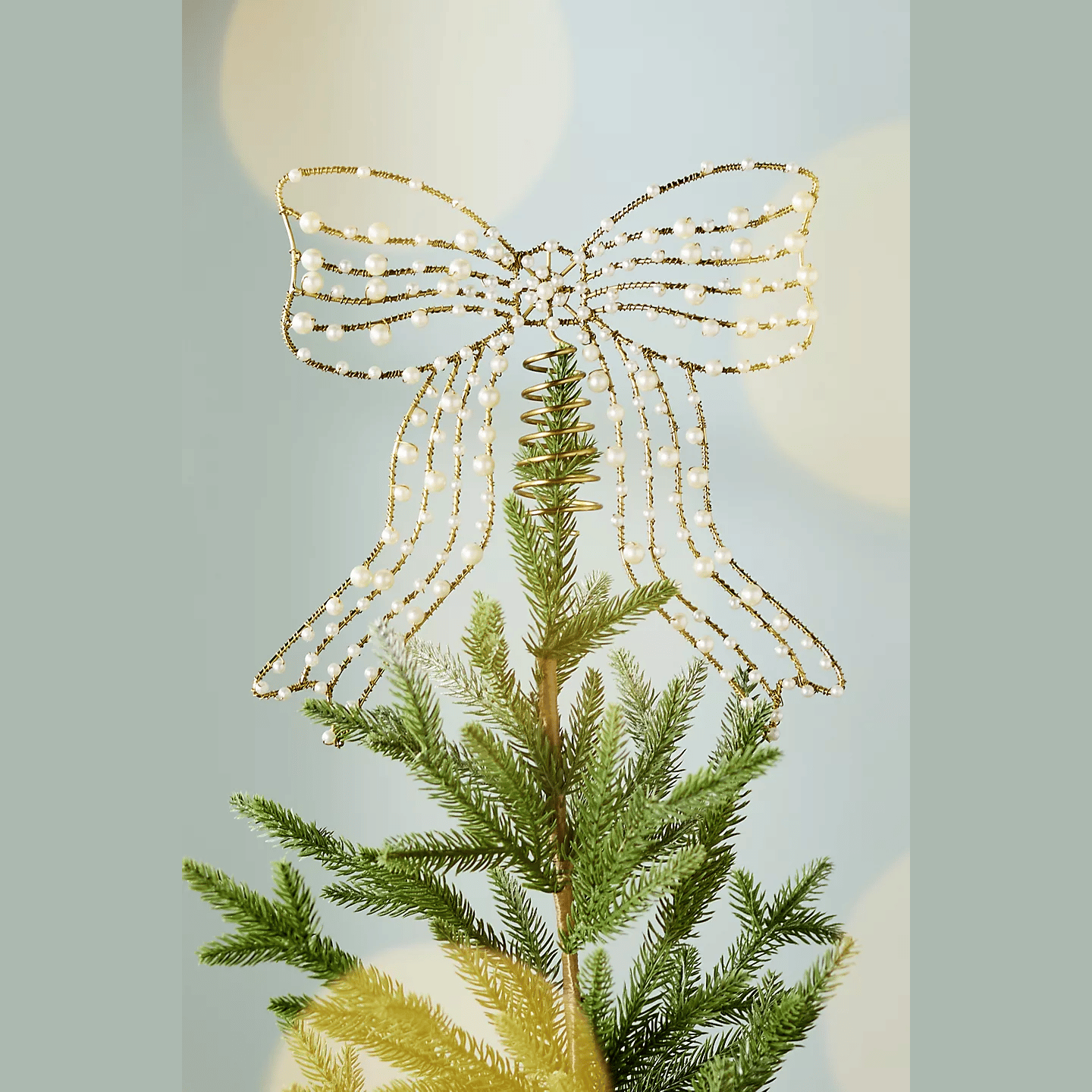 A gold and pearl beaded bow tree topper sits atop a green pine tree, featuring a spiral base for attachment.